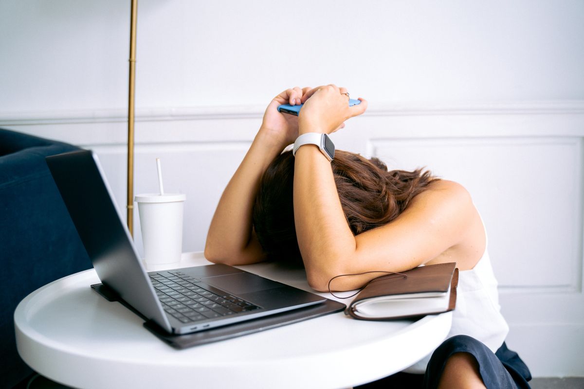 Mulher com a cabeça baixa sobre a mesa, ao lado do notebook, mostrando cansaço e travamento